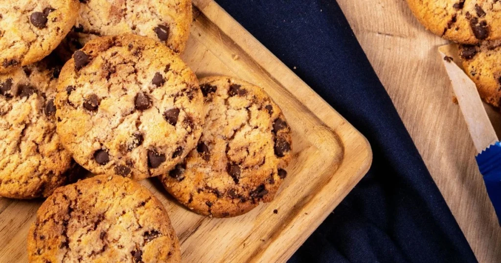 Freshly baked chocolate chip cookies arranged on a wooden serving board with a blue napkin beside it